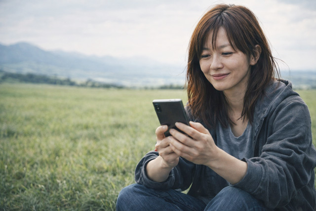 草原に座り笑顔でスマホを見る女性